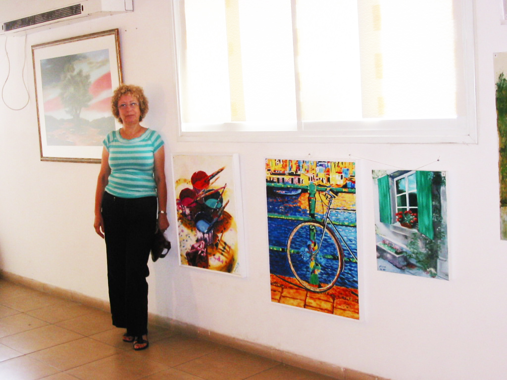 A woman in a turquoise striped top stands beside colorful paintings displayed beneath high windows in a bright exhibition room.