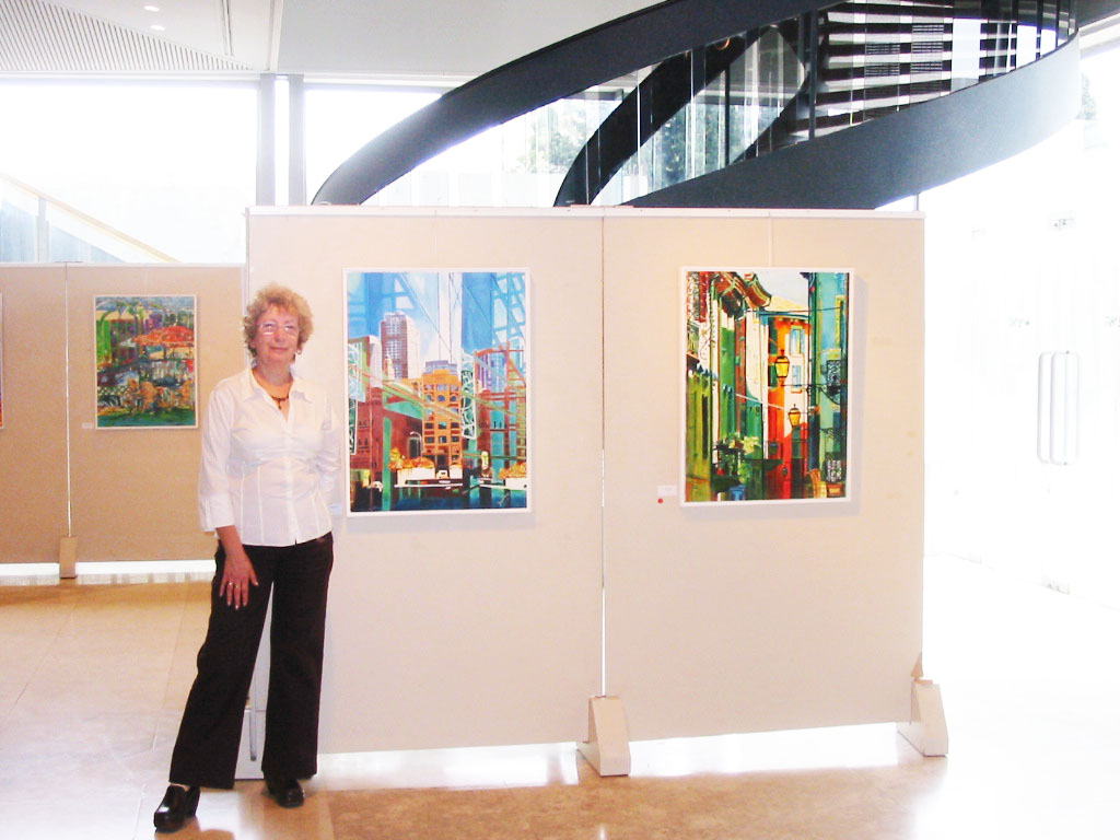 A woman stands beside paintings mounted on pale display panels in a bright foyer beneath a curved staircase.