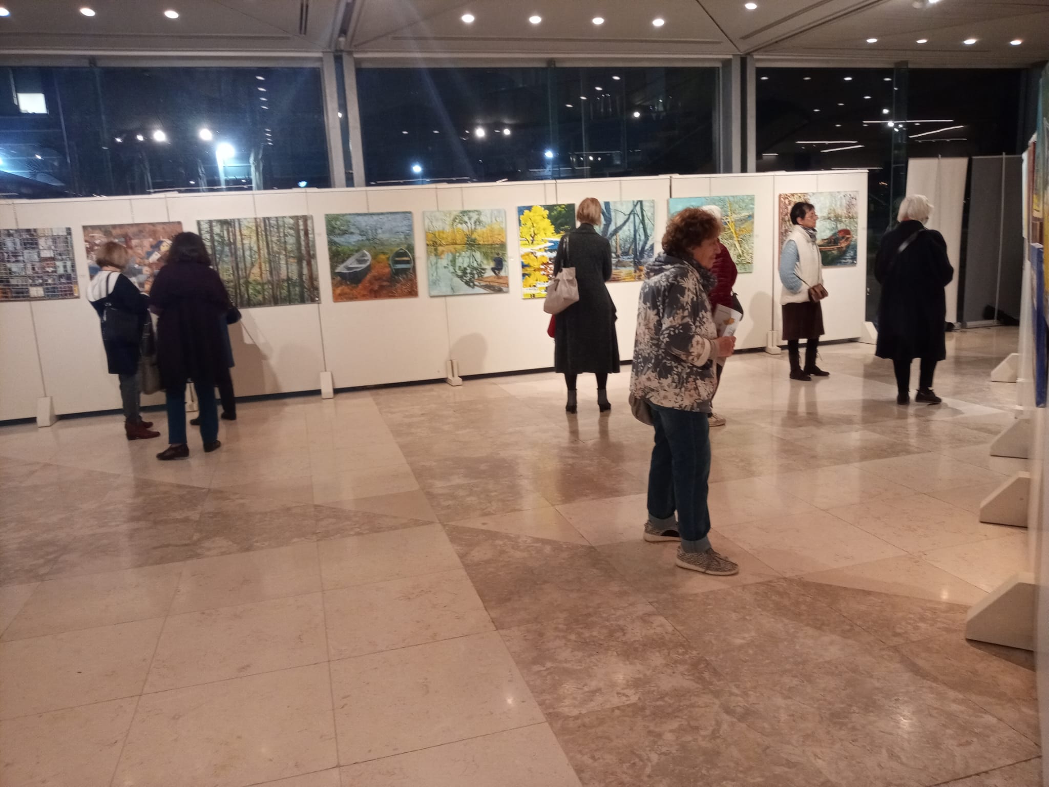 Visitors move between white exhibition panels displaying paintings in a spacious foyer with large dark windows.