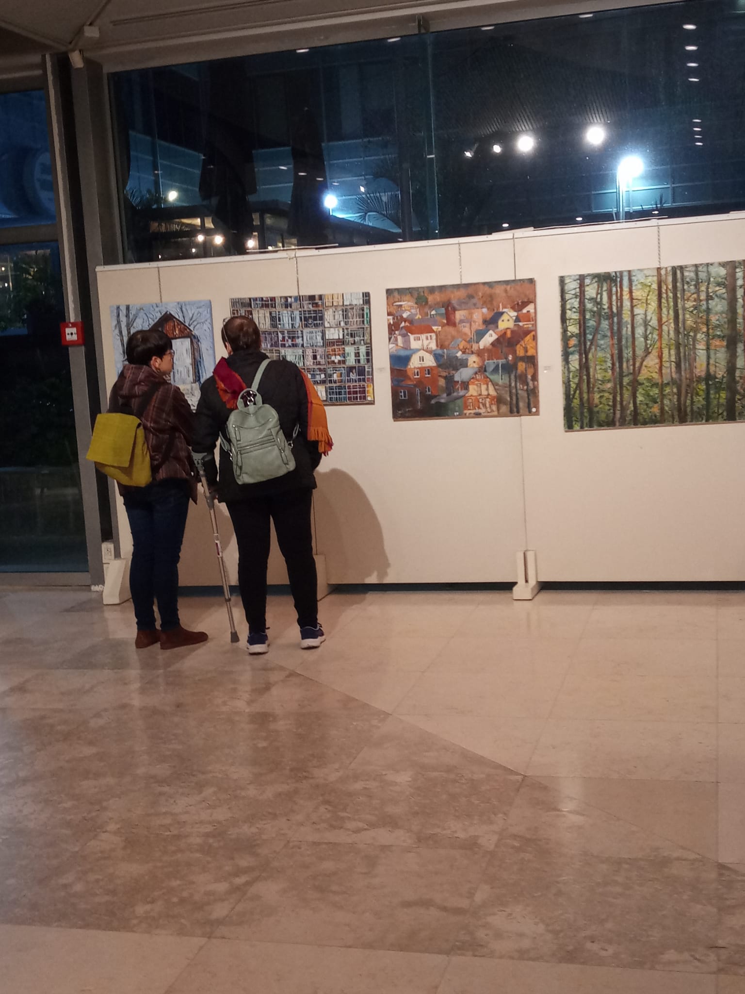 A small group of visitors gathers before paintings on pale display panels beneath the foyer's illuminated ceiling.