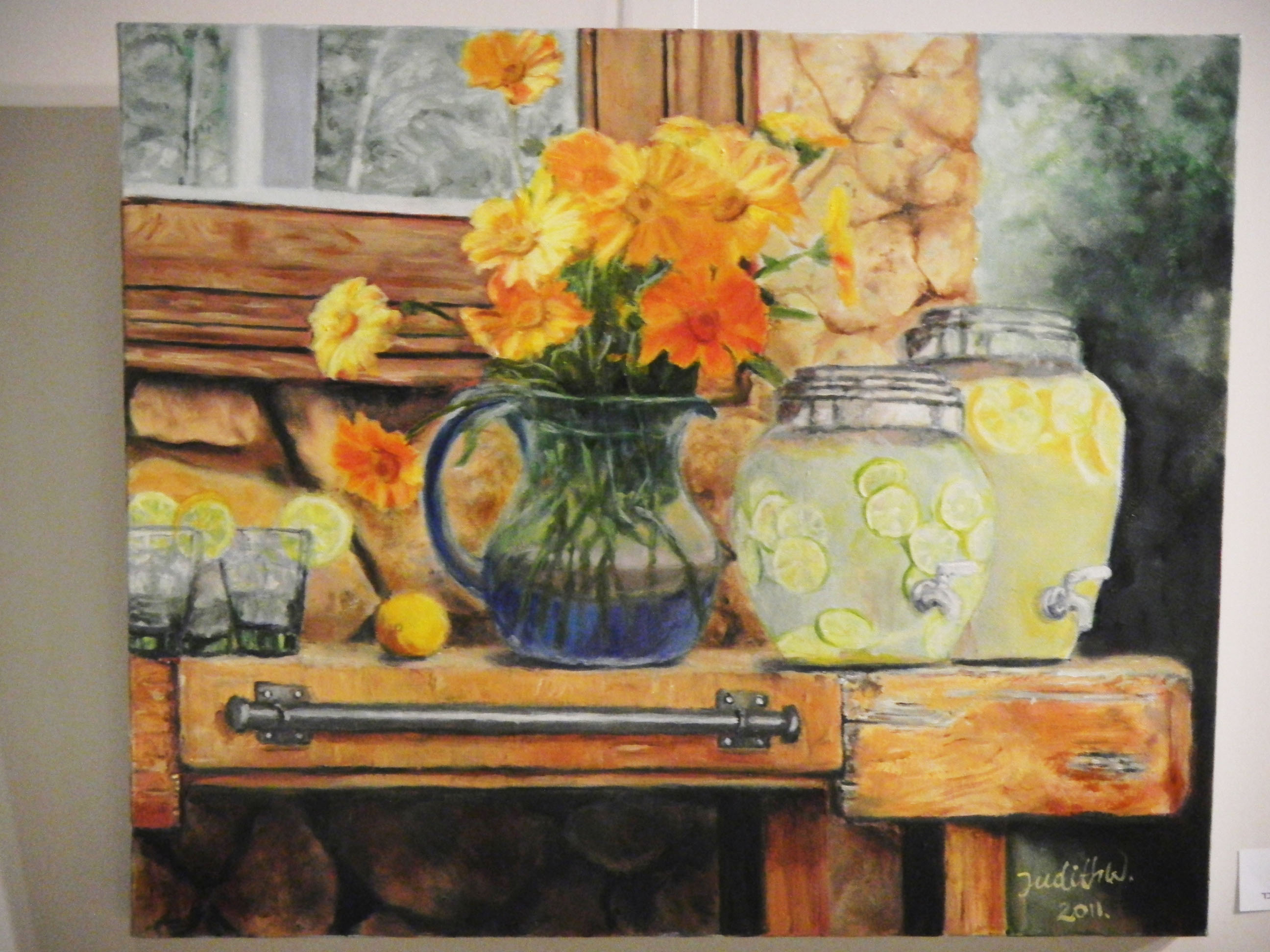 A close view of an exhibited still-life painting shows yellow flowers, a lemonade pitcher and a wooden cabinet within a pale frame.