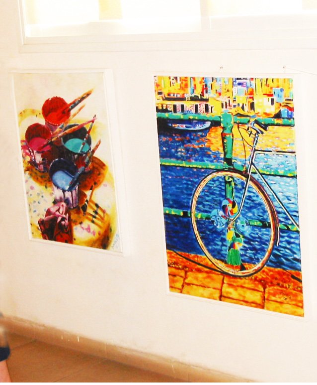 Two paintings of paint pots and a bicycle hang below a bright window on a white exhibition wall.