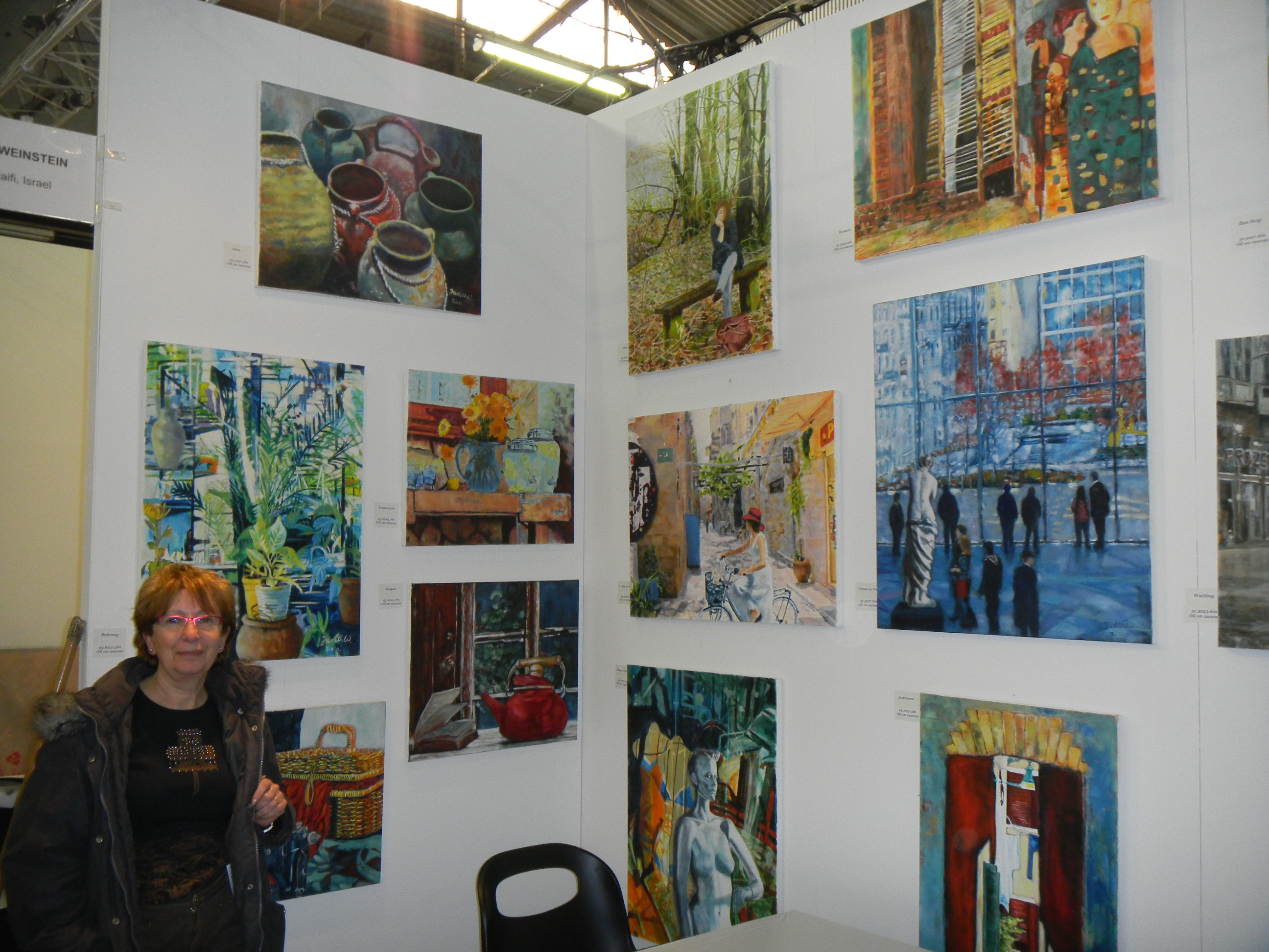 A woman sits inside an ArtExpo booth whose white walls are filled with colorful paintings of plants, streets and people.