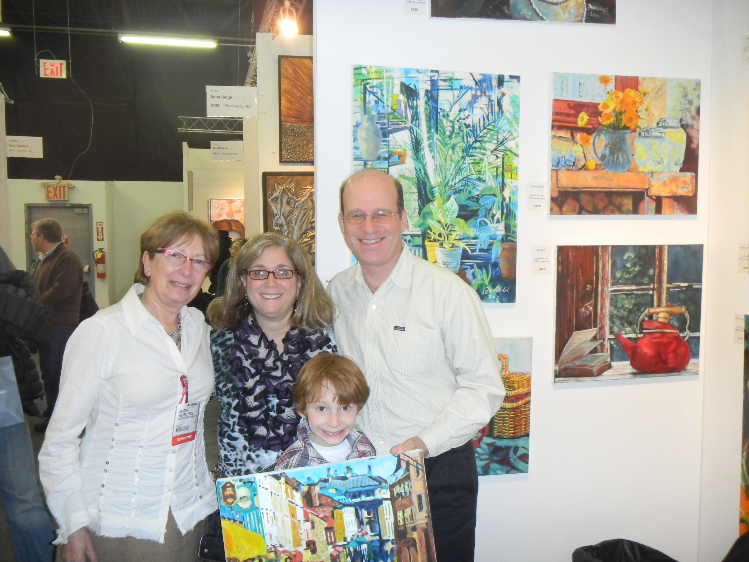 Four people, including a child, pose in front of paintings displayed on the white walls of an ArtExpo booth.