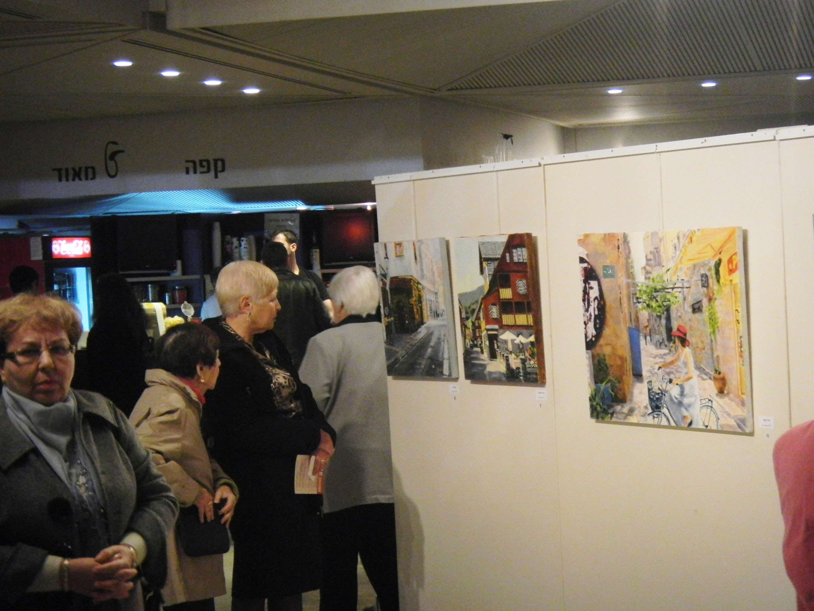 Visitors stand and talk beside a long row of paintings on white display walls in an exhibition hall.