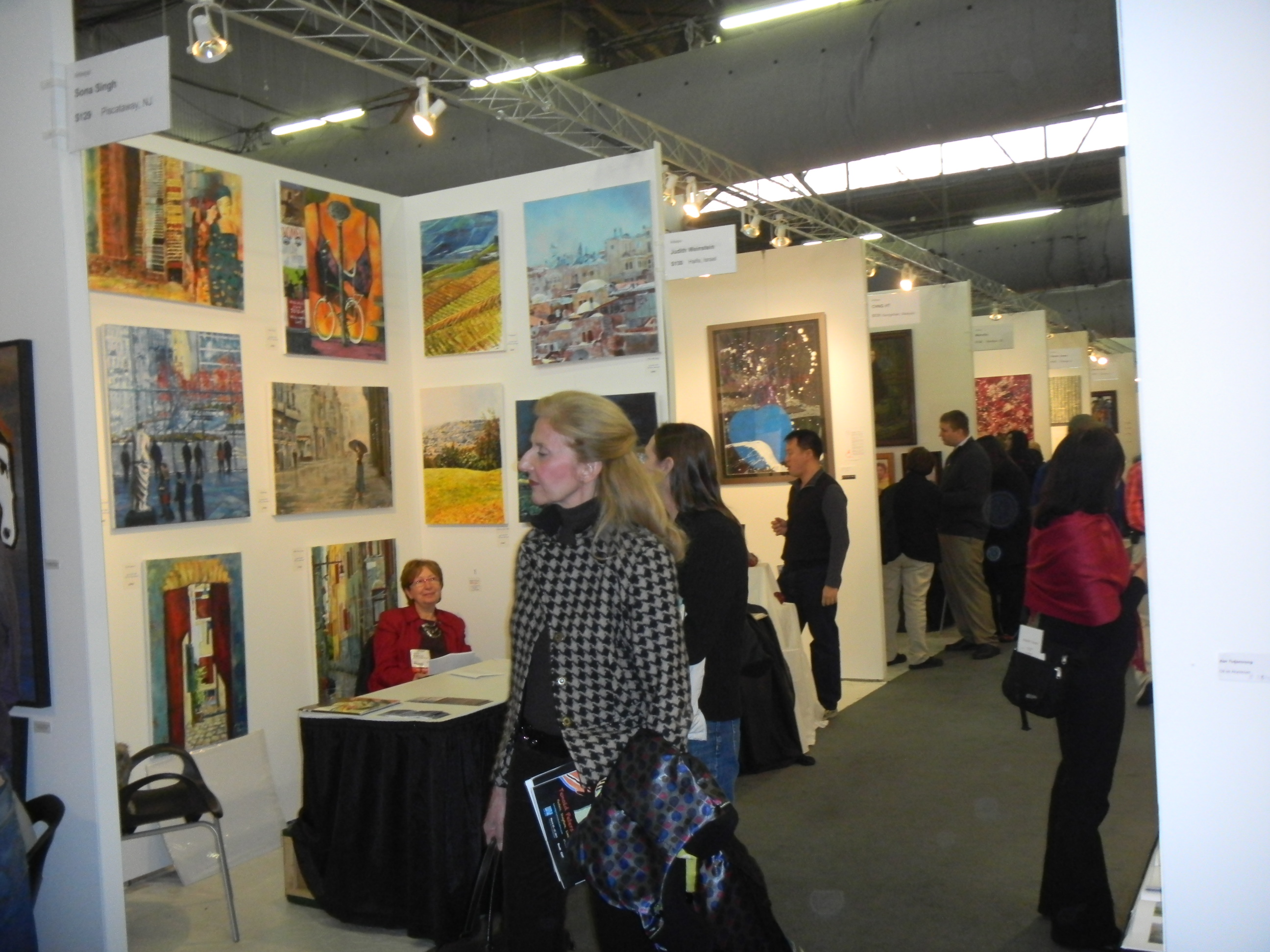 Visitors walk past a densely hung ArtExpo booth, with colorful paintings above a small table and seated exhibitor.