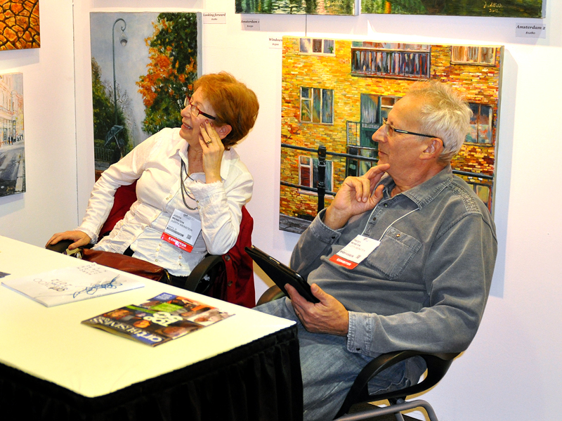 Two people sit at a table with printed material beneath paintings displayed inside an ArtExpo booth.