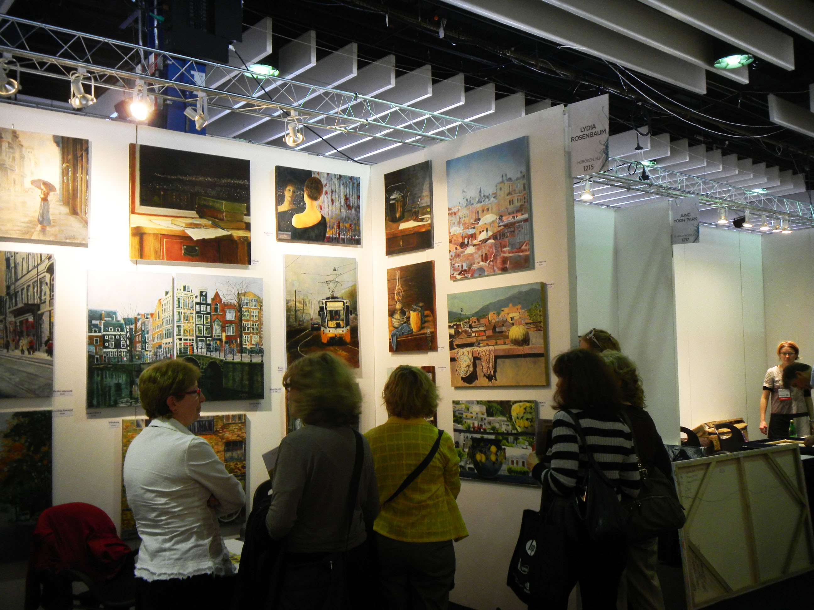 Visitors gather around an ArtExpo booth with paintings hung closely across white walls beneath overhead exhibition lights.