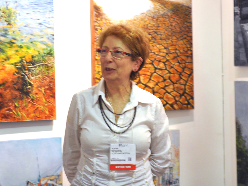 A woman wearing red glasses and an exhibitor badge stands beside two brightly colored paintings on a white ArtExpo booth wall.