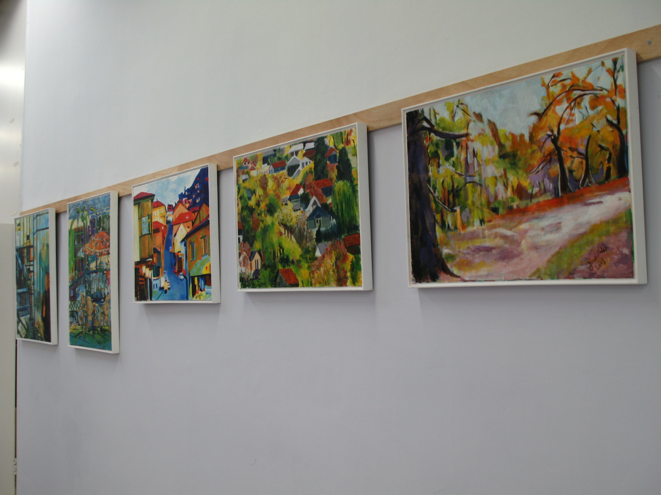 Five framed paintings hang in a row along a white exhibition wall, including landscapes and colorful street scenes.