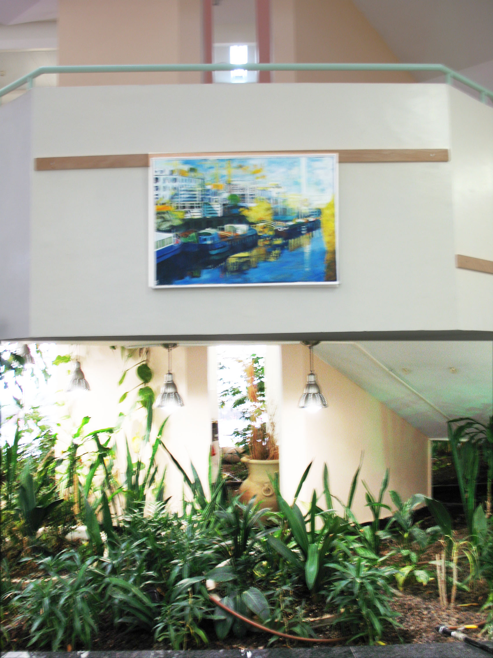 Two views of an exhibition space show a framed harbor painting above a balcony and leafy plants beside a staircase.