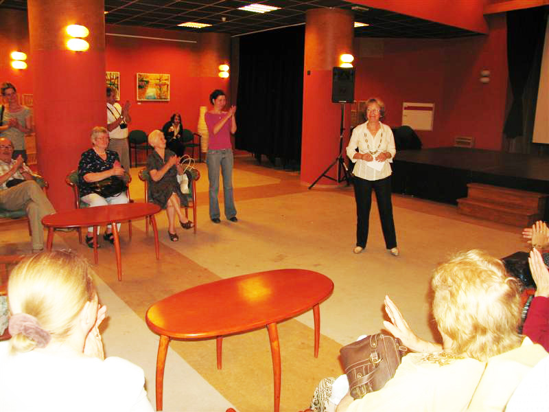 An exhibition speaker addresses seated visitors in a red-walled room, with paintings and small tables around them.