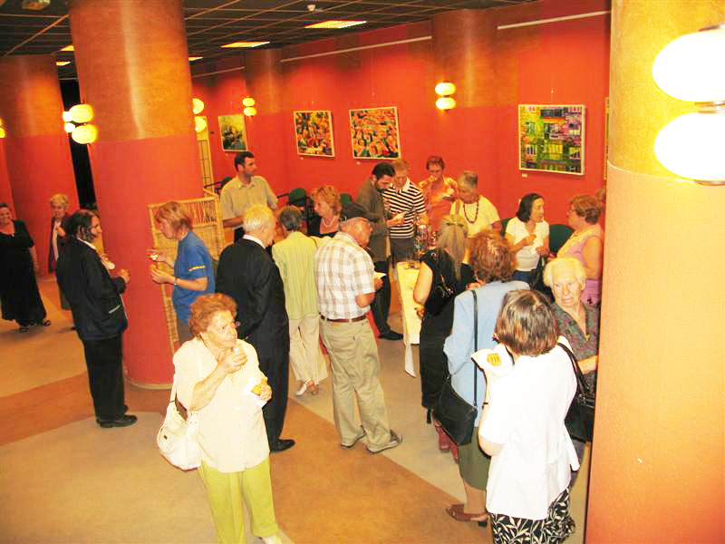 Visitors gather and talk in a red-walled exhibition room, with small framed paintings displayed behind them.