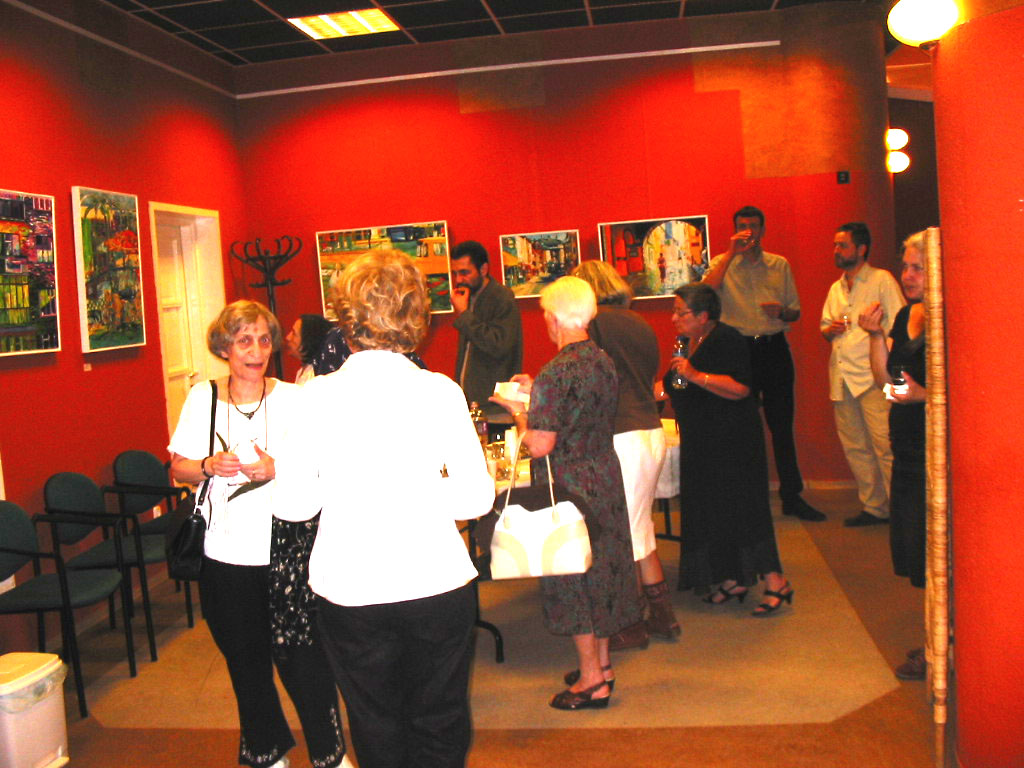 Visitors converse beneath a row of framed paintings in a red gallery room, with chairs lined against the wall.