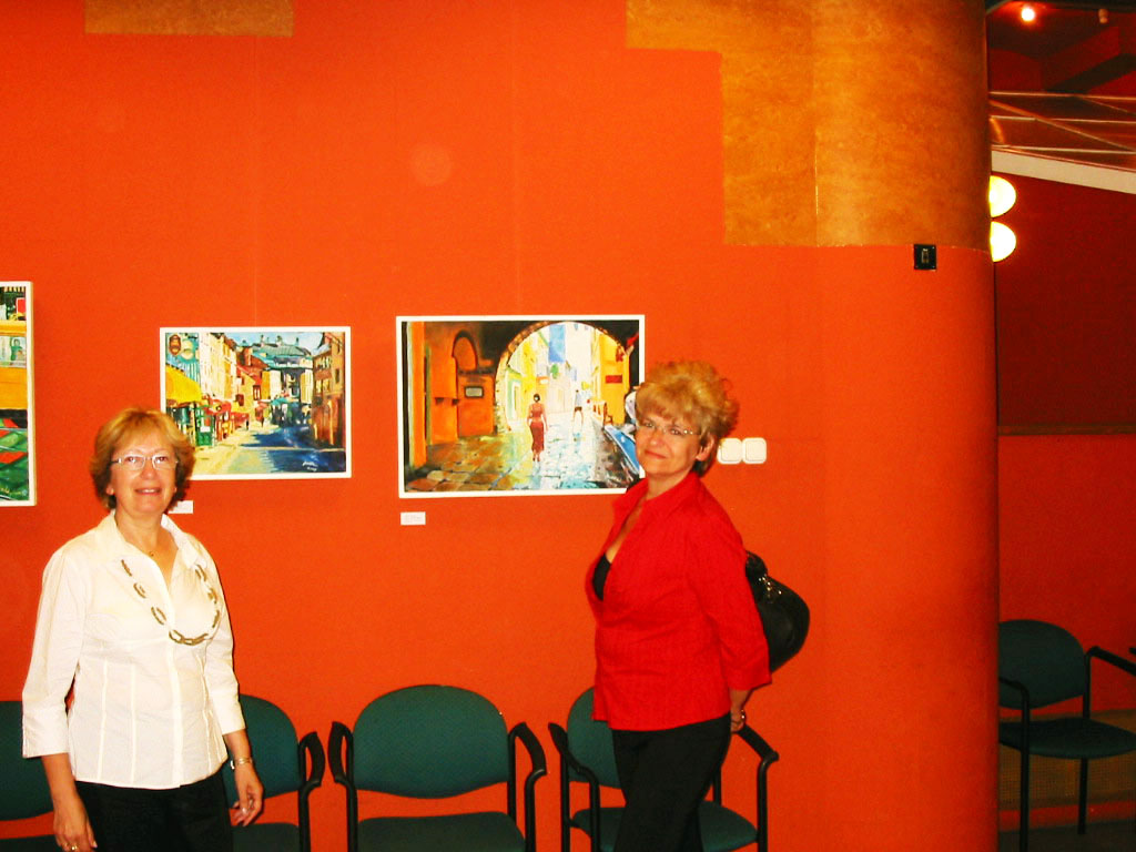 Two women stand beside two framed paintings against a red gallery wall, with chairs lined below.