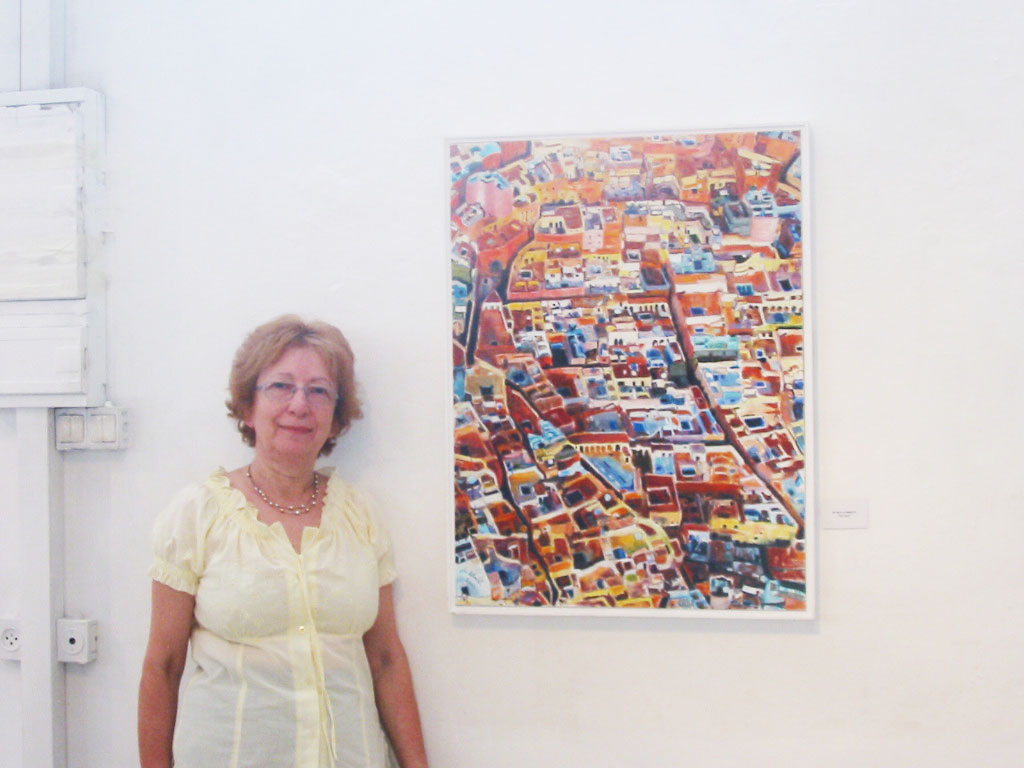 A woman in a pale blouse stands beside a framed, densely colored cityscape on a white wall at the Beit Aba Husi exhibition.