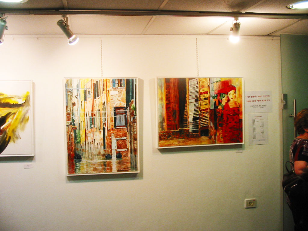 Framed street and city paintings hang on a white exhibition wall beneath spotlights, with an information sheet at the right.