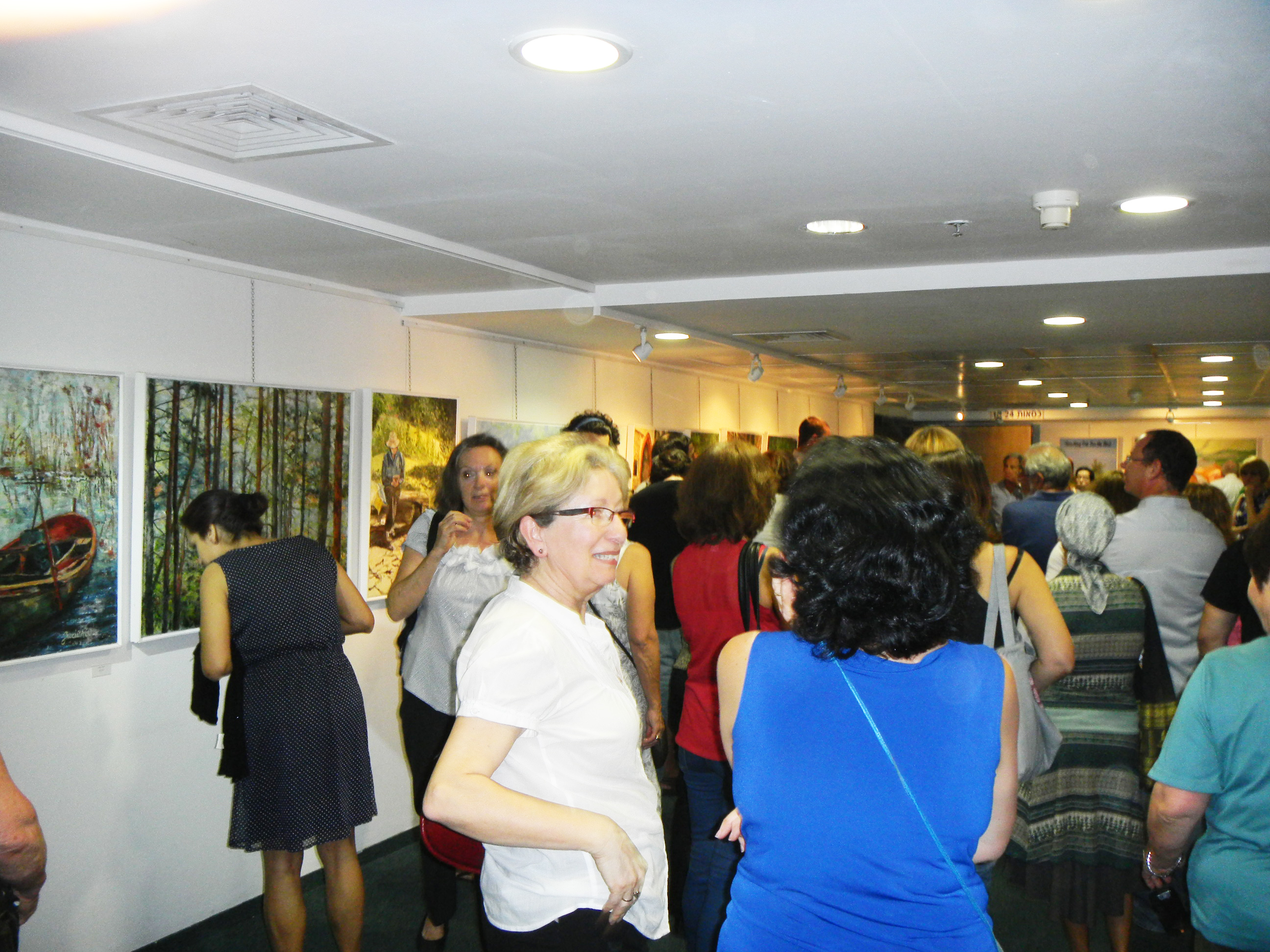 Visitors converse in a crowded exhibition gallery, with framed paintings lining the walls and ceiling spotlights overhead.