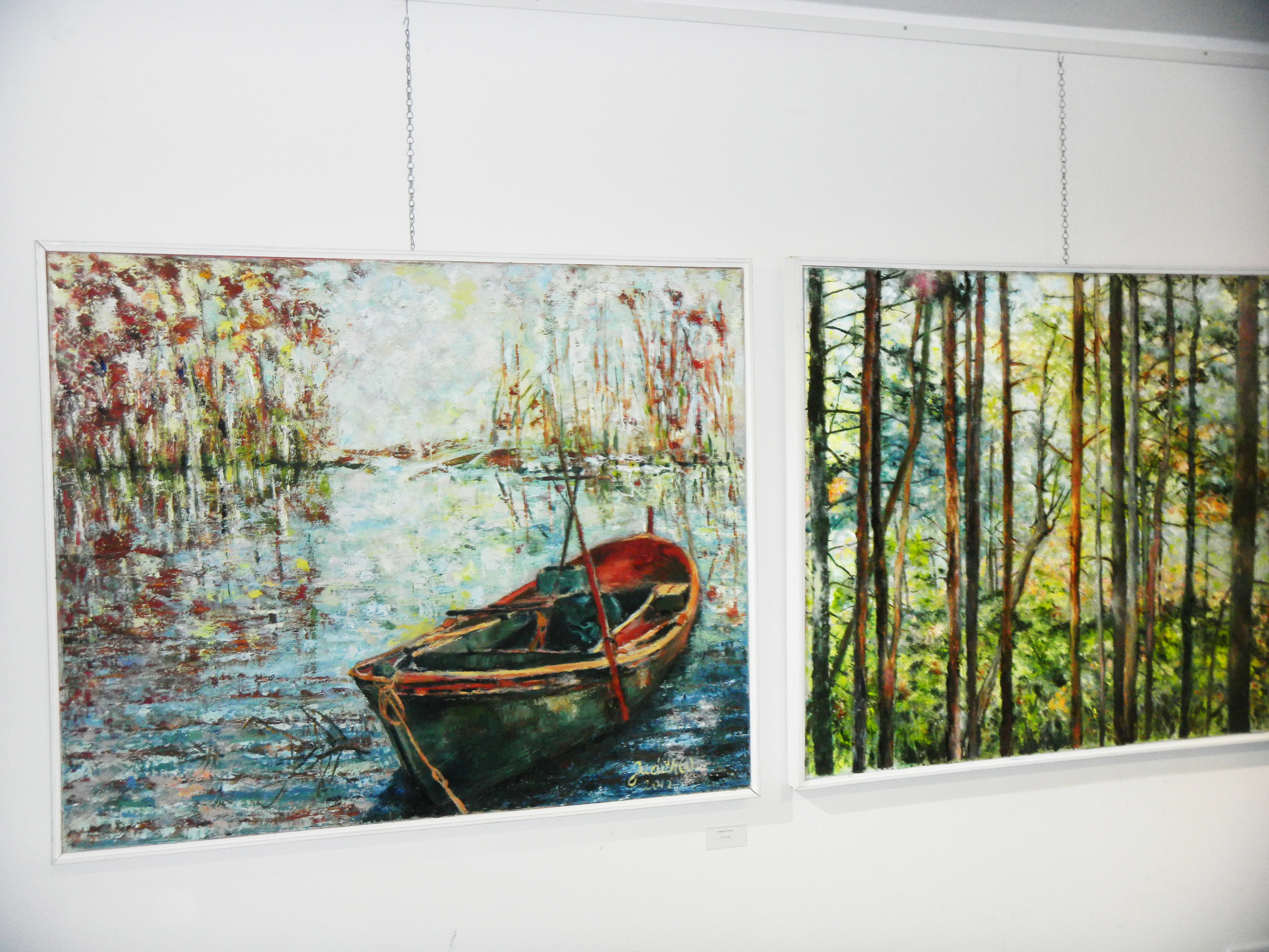Two framed woodland paintings hang side by side: a rowboat beside reeds and tall trees in dappled light.