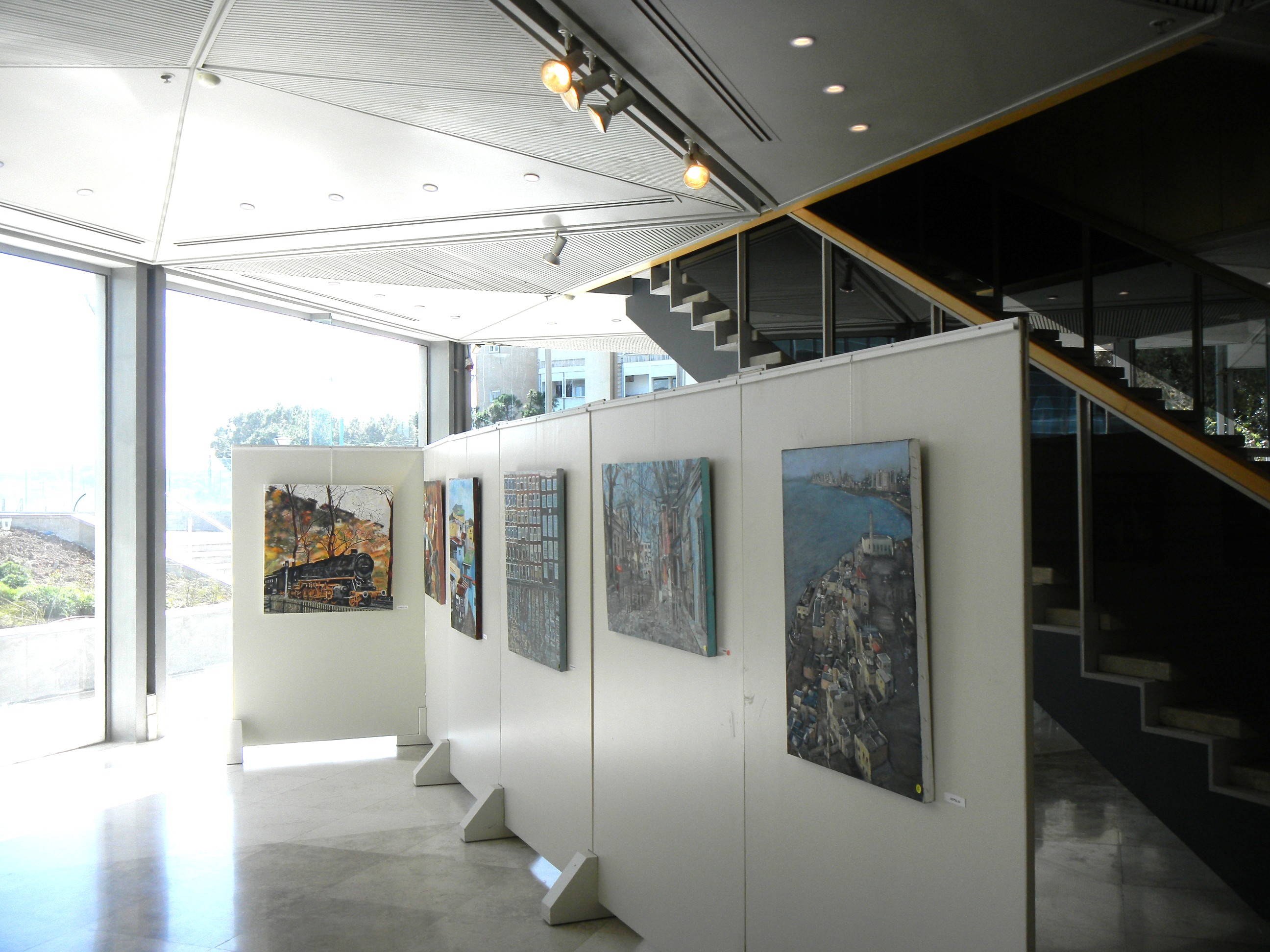 Framed paintings line temporary white exhibition panels beneath a staircase in a bright glass-walled foyer.