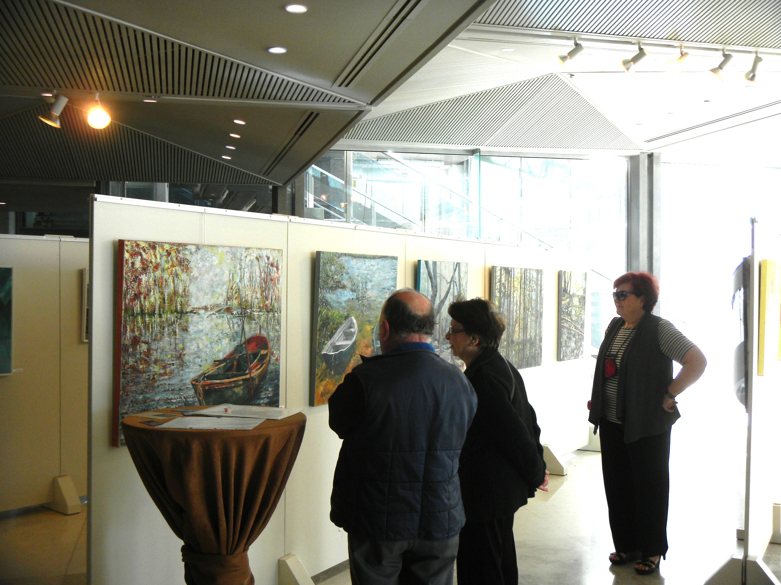 Three visitors discuss paintings beside a tall round table and white display panels beneath gallery spotlights.