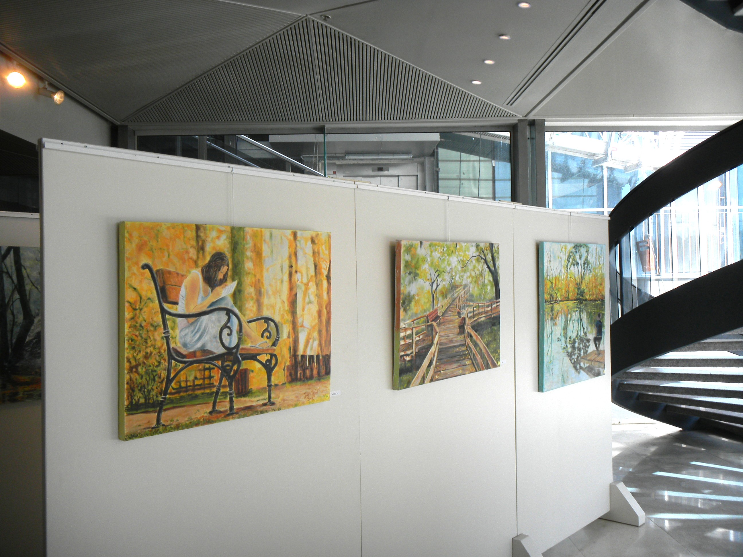 Paintings of a seated reader and woodland scenes hang on white display panels beside a curved staircase.