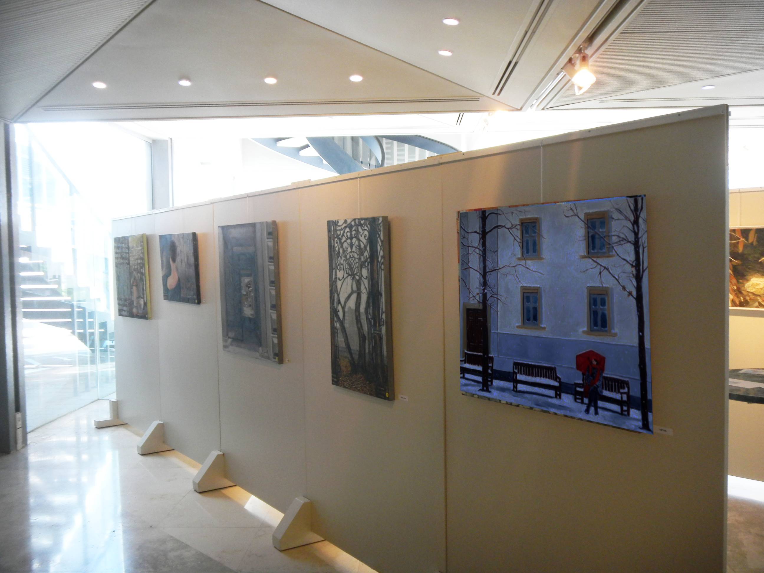 A row of paintings hangs on freestanding white panels, including a snowy street scene, in a bright gallery foyer.