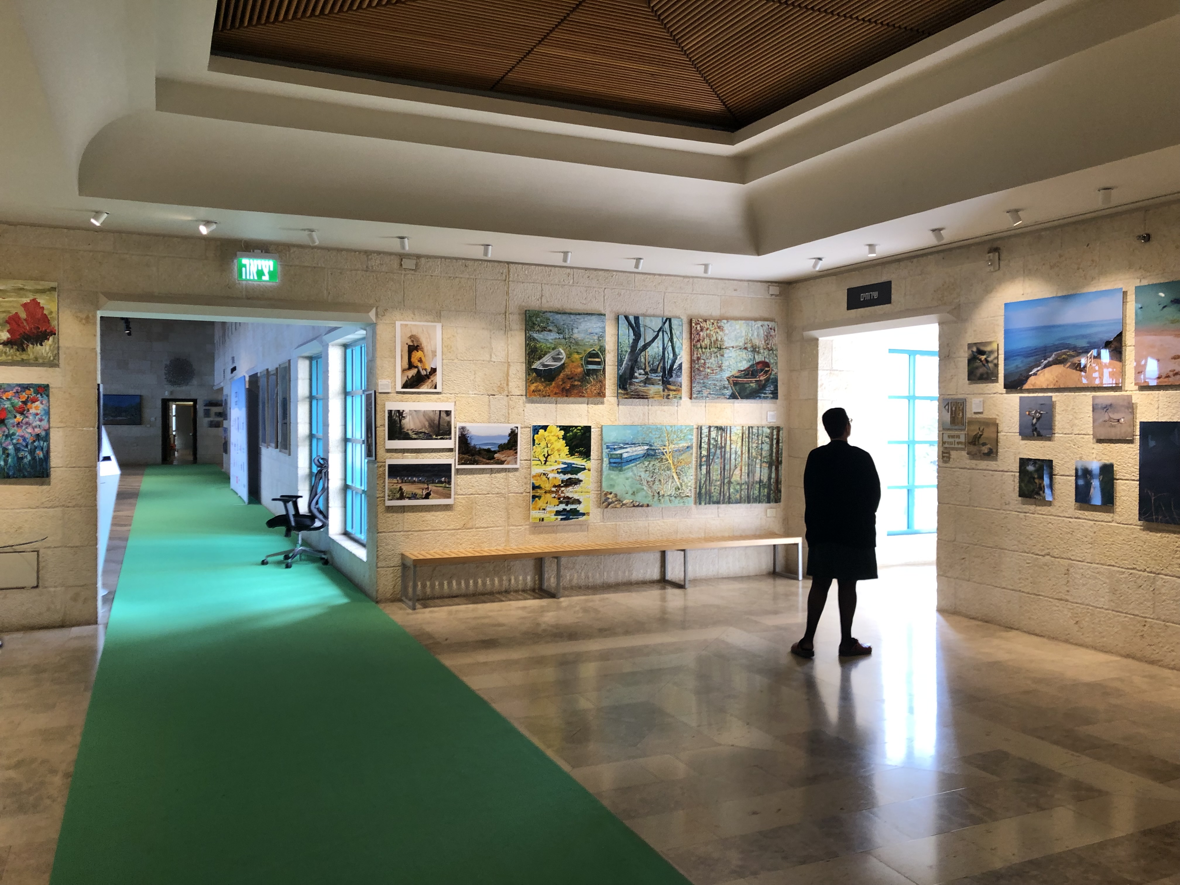 A visitor walks through a stone-walled exhibition foyer, with paintings around the room and a green carpet along the corridor.