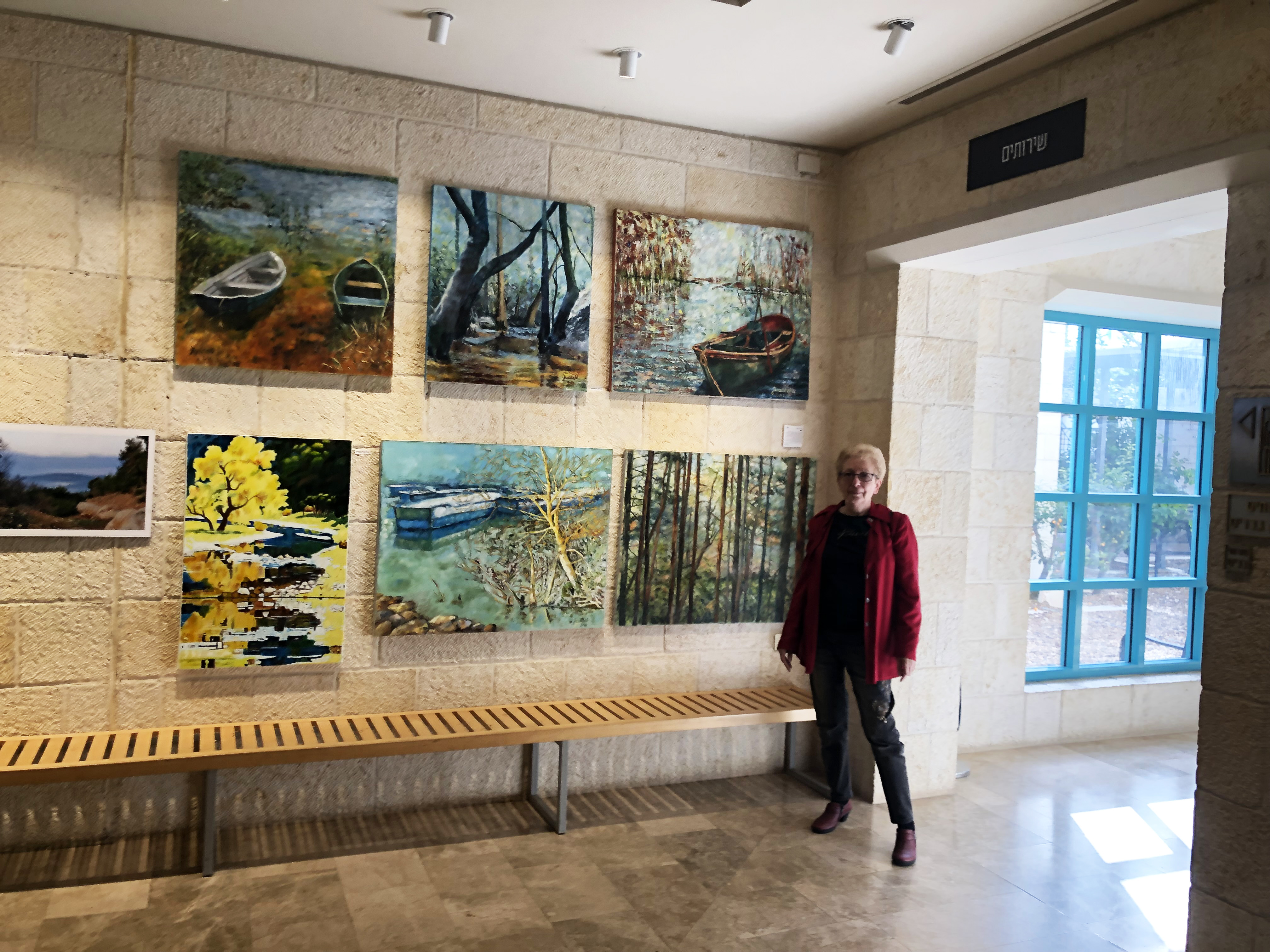 A woman in a red jacket stands beside landscape paintings displayed above a wooden bench on a pale stone wall.