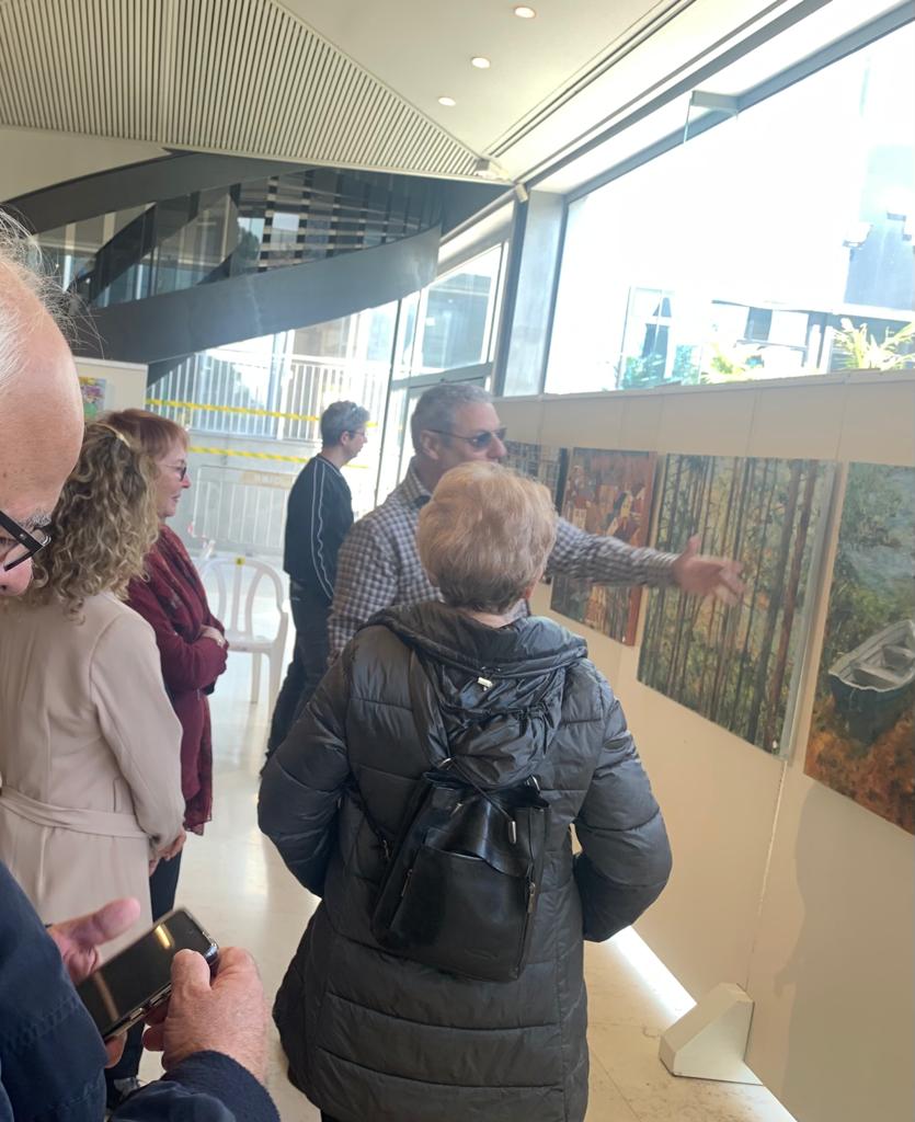 A visitor gestures toward woodland paintings while a small group listens beside an exhibition panel in a bright foyer.