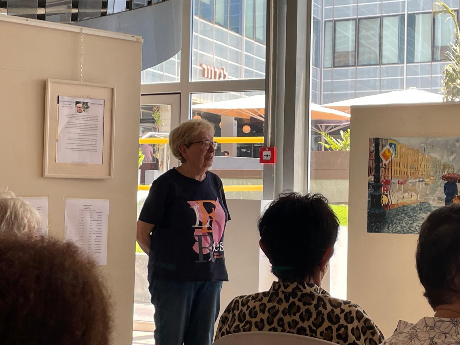 A woman wearing a black graphic T-shirt addresses seated visitors beside an exhibition panel in a bright glass-walled foyer.
