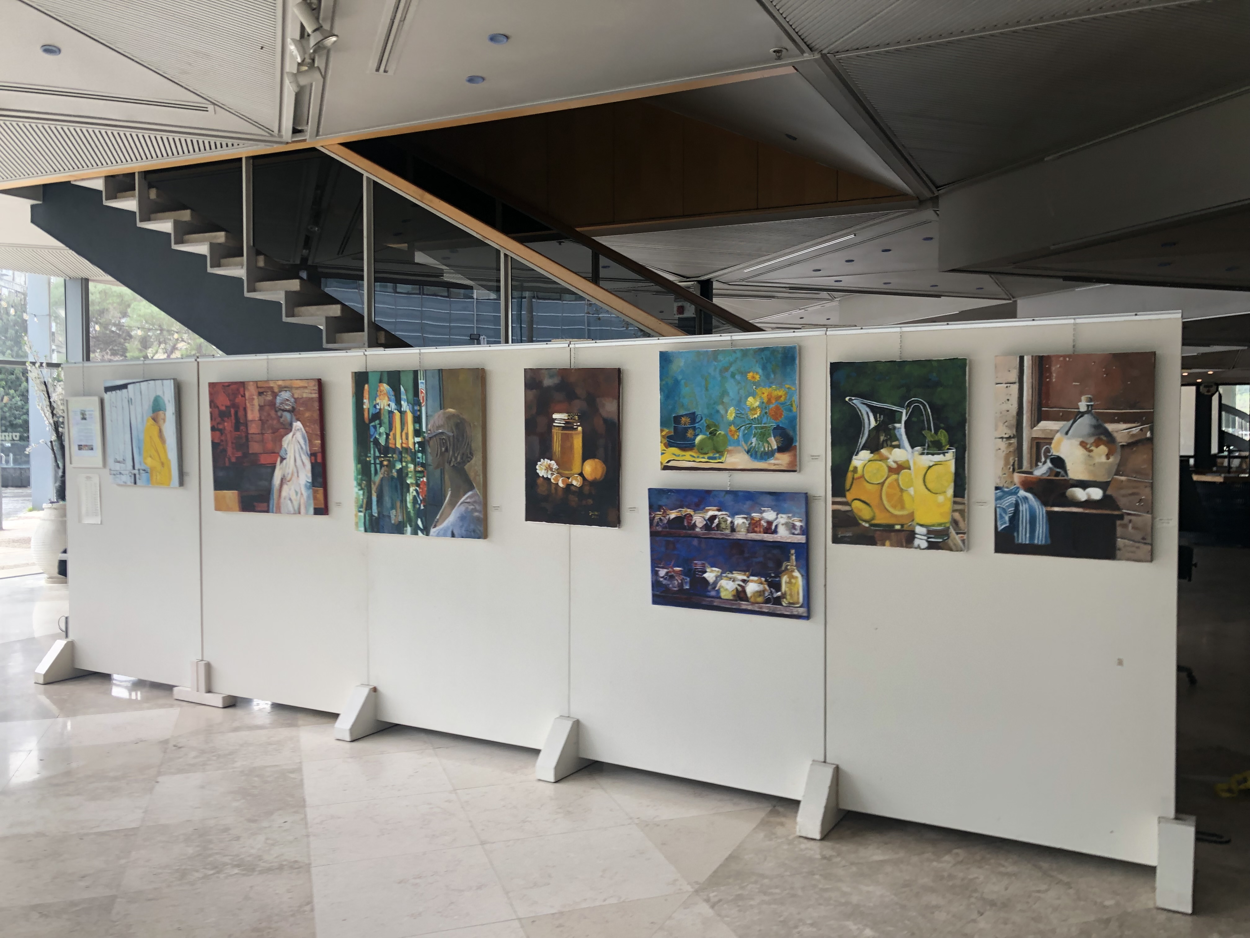 Paintings of streets, flowers and still-life arrangements hang across freestanding white panels beneath a broad gallery staircase.