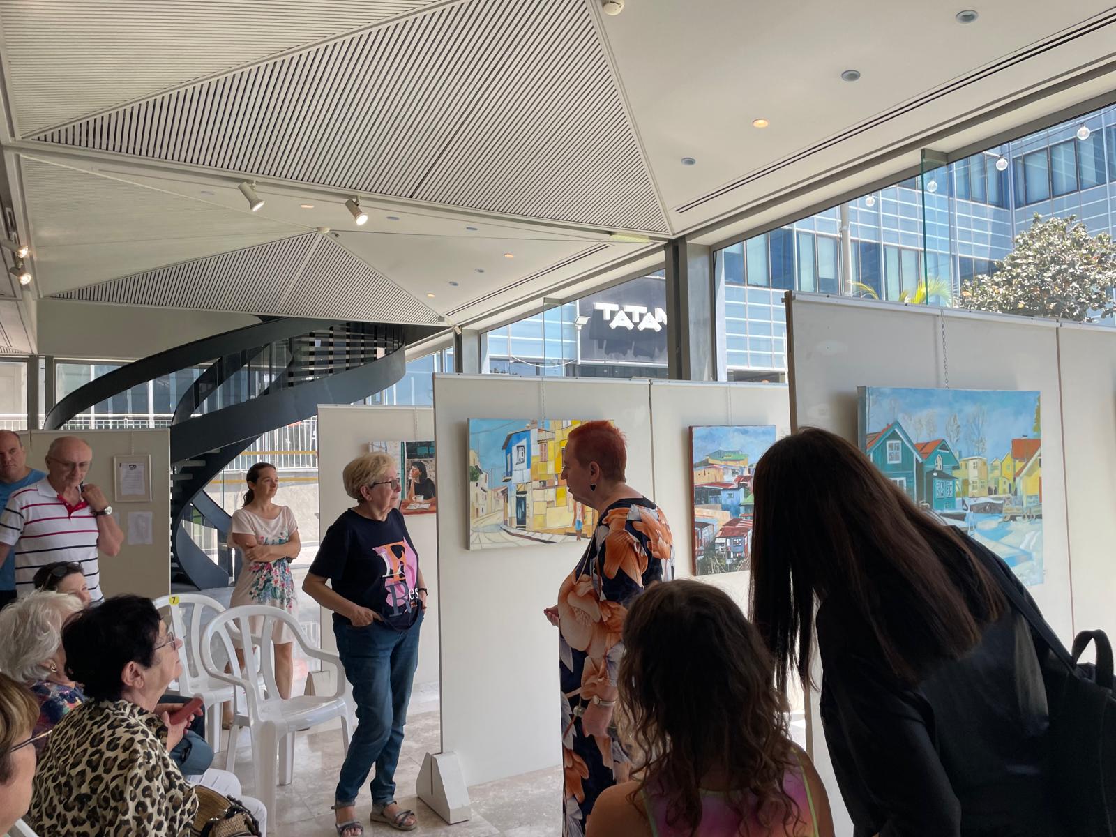 Visitors gather around a speaker beside white exhibition panels, with a harbor painting and large windows behind them.