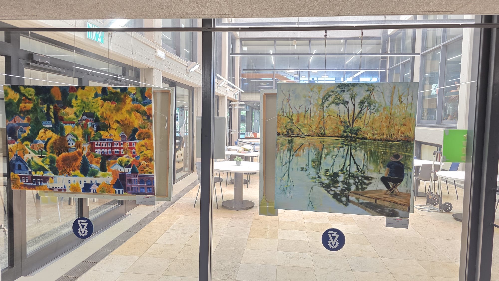 Colorful hillside and lake paintings hang against glass panels in a sunlit corridor, with doors and further artwork beyond.