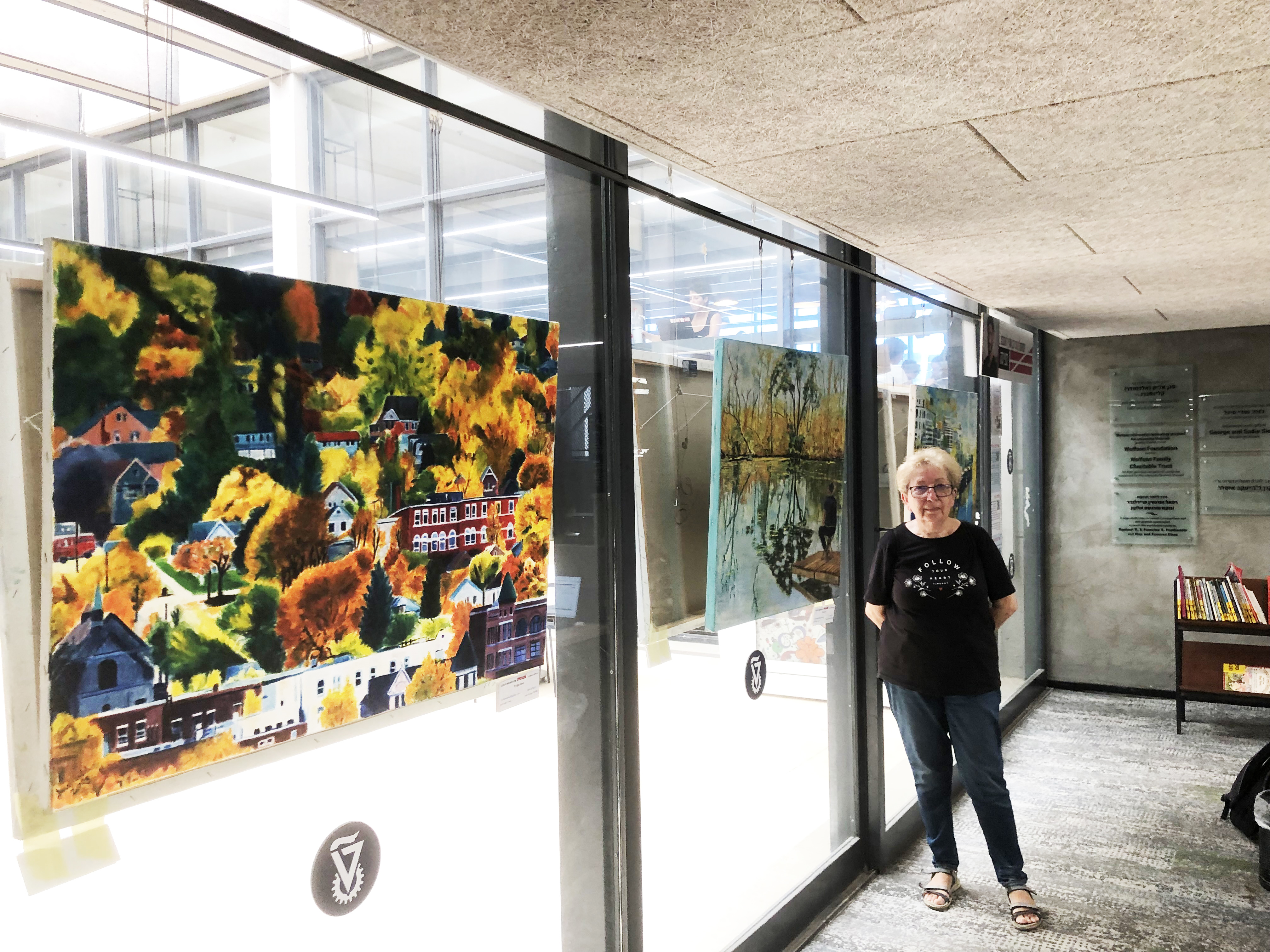 A woman stands beside paintings hung along a glass corridor, including a colorful hillside and a calm lakeside scene.